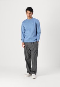 Sudadera azul claro con cuello redondo, puños acanalados y un pequeño logo; combinada con pantalones grises con pliegues y zapatillas blancas.