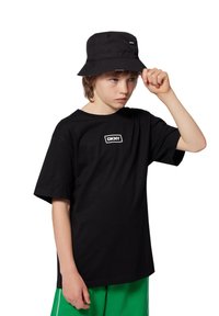 T-shirt en coton noir à manches courtes avec un logo rectangulaire blanc. Accompagné d'un chapeau de type bucket noir avec un logo assorti. Short vert.