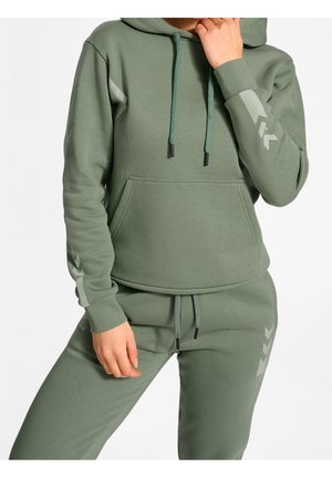 Person trägt olivgrünen Hoodie und passende Jogginghose mit hellgrauen Chevron-Mustern an Ärmeln und Hosenbein.