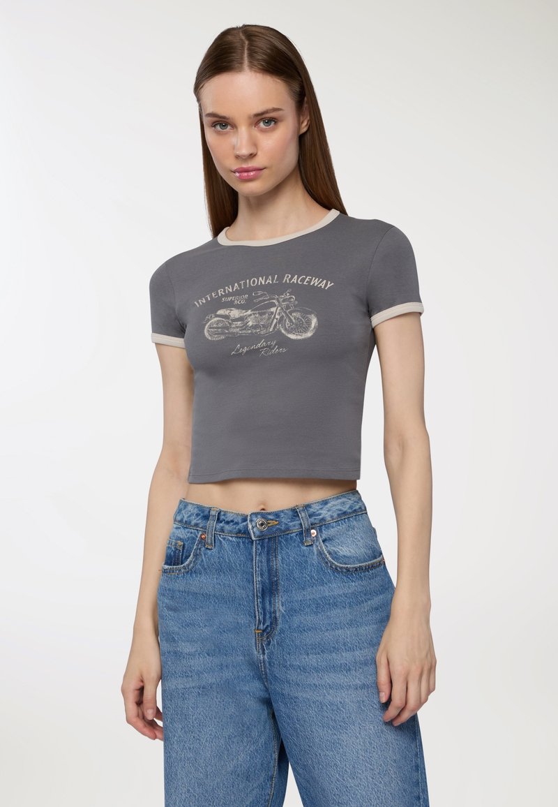 Młoda kobieta z prostymi brązowymi włosami, ubrana w szary krótki T-shirt z motywem motocykla oraz dżinsy o wysokim stanie w niebieskim kolorze, na jednolitym tle.