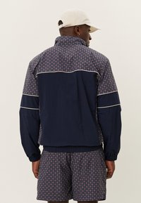 Námornícky vetrovka so geometrickým vzorom, vysokým golierom, elastickými manžetami a pásom. Obsahuje kontrastne biele lemovanie. V kombinácii s zodpovedajúcimi šortkami.