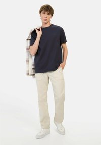 Marineblauwe katoenen t-shirt met ronde hals, gecombineerd met beige katoenen broek. Model houdt een geruit jasje over één schouder. Witte sneakers zichtbaar.