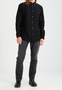 Zwart overhemd met lange mouwen en een button-down kraag, zilveren knopen en een borstzak; gecombineerd met donkergrijze jeans en zwarte schoenen.