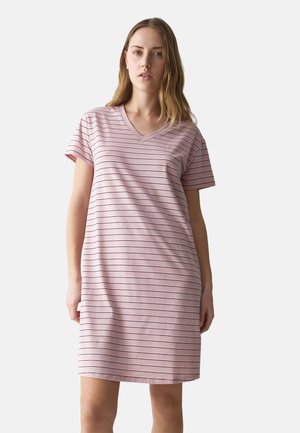 Frau trägt ein pinkfarbenes kurzärmeliges Kleid mit dünnen schwarzen horizontalen Streifen, steht vor einem schlichten hellen Hintergrund.