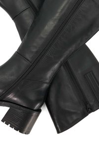 Black leather knee-high boots feature a pronounced heel, side zippers, and a smooth texture with subtle seams. The design is sleek and modern.