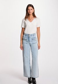 Mujer con una blusa blanca de manga corta con detalles de encaje, jeans de pierna ancha de cintura alta en un tono azul claro con bolsillos frontales, y botas de tacón negras.