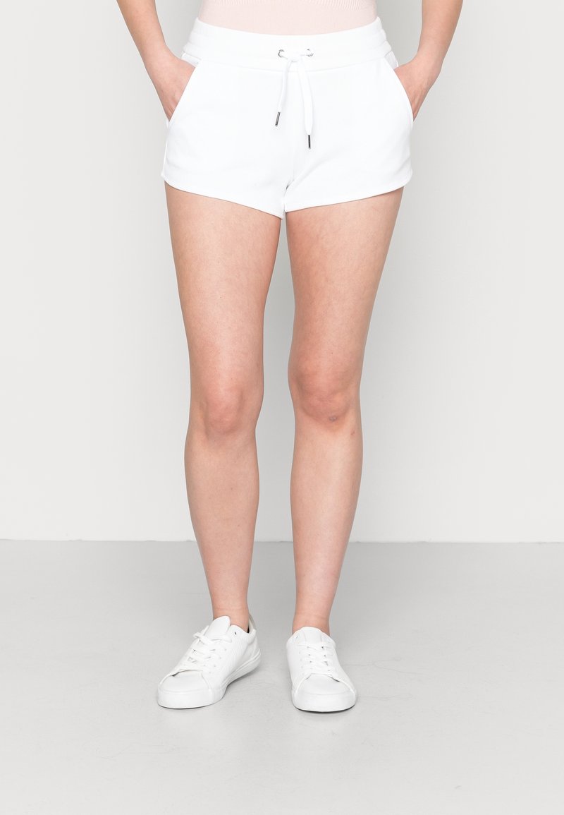 Personne portant un short blanc à cordon de serrage avec les mains dans les poches et des baskets blanches, debout sur un sol gris clair devant un fond uni.
