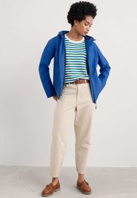 Mėlyna gobtuvinė striukė užmesta ant dryžuoto žaliai baltos spalvos megztinio, béžinės kelnės ir rudų odinių batų. Lygi medžiaga ir laisvas kirpimas.