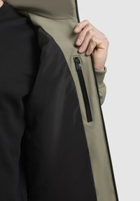 Khaki kabát sima külső felülettel, fekete bélésbel és oldalsó cipzáros zsebbel. Az anyag tartósnak és vízállónak tűnik.