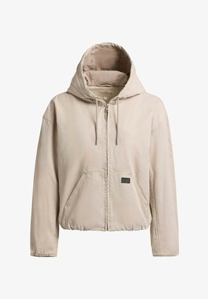 Veste à capuche beige zippée avec cordons de serrage et poches avant, ourlet froncé, et une petite étiquette noire sur la poche.