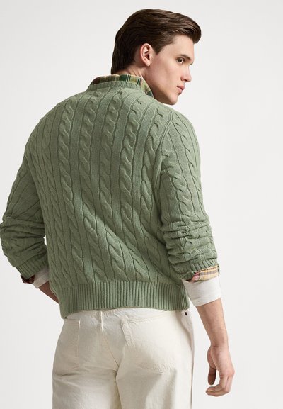 Green cable-knit sweater with a ribbed hem and cuffs, layered over a checkered shirt. The fabric has a textured appearance.