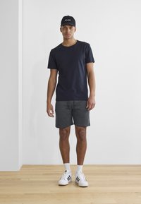 Marineblaues Baumwoll-T-Shirt, graue Shorts und weiße Sneakers mit schwarzen Streifen. Das Model steht vor einer schlichten weißen Wand auf Holzboden.