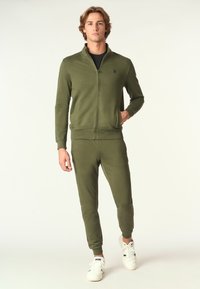 Felpa verde oliva con zip e pantaloni jogger abbinati, realizzati in tessuto morbido. Presenta polsini e vita a coste, abbinati a sneakers bianche.