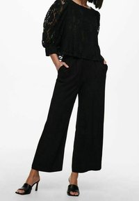 Zwarte kanten top met opgeblazen mouwen, gecombineerd met zwarte broek met wijde pijpen. De outfit wordt aangevuld met zwarte open-toe sandalen met hak.