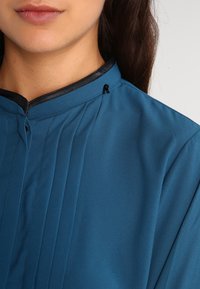 Blusa teal realizzata in tessuto morbido, caratterizzata da un design plissettato, colletto nero testurizzato e un piccolo tag nero vicino allo scollo.