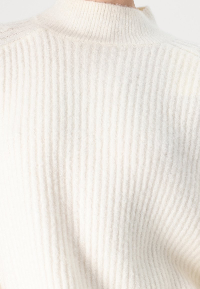 Maglione bianco a coste con colletto alto e tessuto morbido e testurizzato con una leggera lucentezza. Il design enfatizza calore e comfort.