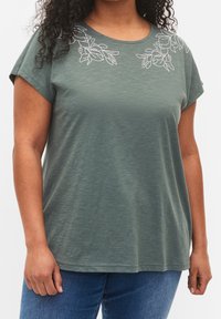 Kvinna i grön kortärmad topp med vit blommönsterlinje vid halsringningen och blå jeans, som står framför en vit bakgrund.