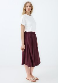T-shirt blanc à manches courtes associé à une jupe midi bordeaux avec une taille élastique, une silhouette fluide et des sandales beiges.