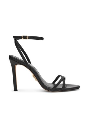 Sandale noire à talon haut avec des fines brides croisées sur les orteils et une bride à la cheville avec une boucle dorée, présentée sur fond blanc.