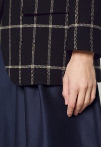 Ruka odpočívala po boku, oblečená v černobílé kostkované bundě s rukávem přes tmavě modrou plisovanou sukni.