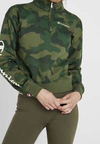 Pullover camouflage verde con colletto alto e cerniera a metà. Presenta il logo Champion sul petto e sulle maniche. Abbinato a leggings oliva.