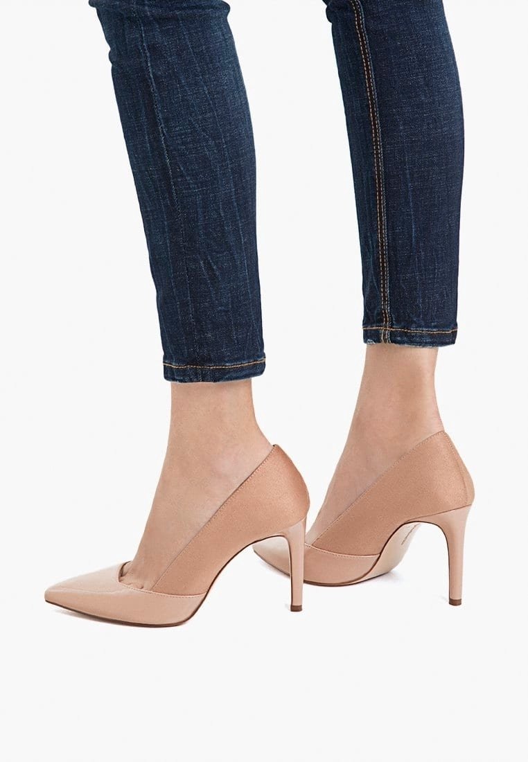 Jambes en jean skinny foncé portant des escarpins nude à bout pointu et talons stilettos sur un fond blanc.