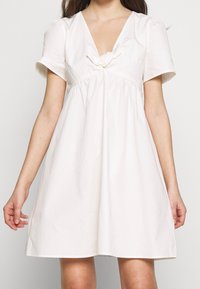 Vit bomulls klänning med korta ärmar, V-ringning och samlad midja. Har en knutdetalj vid bröstet och en utställd nederkant.