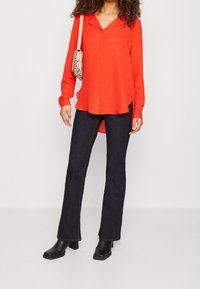 Blouse rouge à manches longues avec un col en V, associée à un jean évasé en denim foncé et des bottines à talons noires. Comprend un petit sac à main à motif.