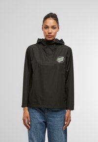 Black anorak jacket featuring a hood, front pocket, and a decorative logo. Made from water-resistant material with a smooth texture.