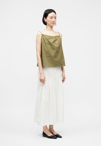 Γυναίκα με κοντά μαύρα μαλλιά που φοράει αμάνικο τοπ ελιάς, μακριά λευκή φούστα και μαύρα μυτερά παπούτσια, στέκεται μπροστά σε λευκό φόντο.