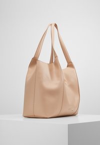 Beige texturerad läder-handväska med dubbla axelremmar, rektangulär botten och subtil logotyp på framsidan, uppställd på vit pelare.