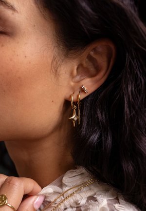 Boucles d'oreilles en or avec un pendentif pointu et un petit accent en diamant, associées à une boucle d'oreille en argent en forme d'étoile. De longs cheveux bruns tombent à proximité.