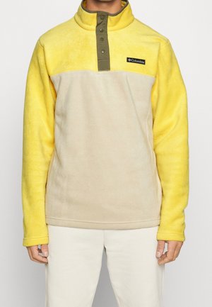 Mężczyzna ubrany w żółto-beżowy pulower polarowy Columbia z kołnierzem na guziki i białe spodnie, stojący na tle gładkiego tła.