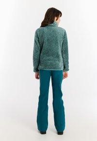 Osoba u tirkiznoj flis jakni i tirkiznim hlačama stoji okrenuta leđima prema bijeloj pozadini.