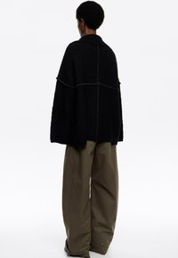 Osoba ubrana w oversize'owy czarny sweter dzianinowy i szerokie oliwkowe spodnie, stojąca tyłem na tle białym.