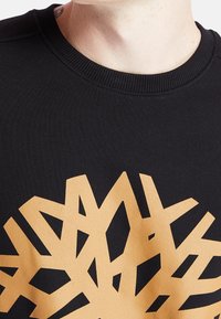Sudadera negra con cuello redondo, que presenta un gran gráfico abstracto dorado en un patrón geométrico en la parte frontal. Textura suave.