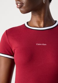 Close-up of a person wearing a red ribbed Calvin Klein t-shirt with white and black trim on the collar and sleeves, silver hoop earring visible.