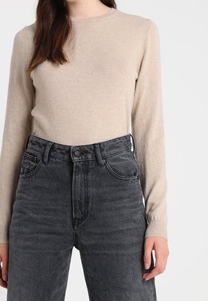 Femme portant un pull beige à manches longues rentré dans un jean en denim noir taille haute avec poches avant et fermeture par boutons.