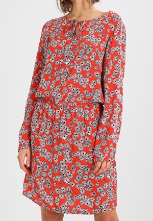 Femme portant une robe rouge à manches longues avec un motif floral de marguerites blanches et noires et un petit nœud à l'avant au niveau du col.