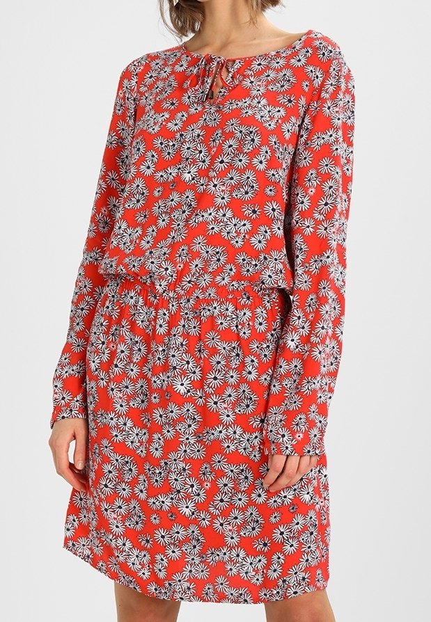 Femme portant une robe rouge à manches longues avec un motif floral de marguerites blanches et noires et un petit nœud à l'avant au niveau du col.