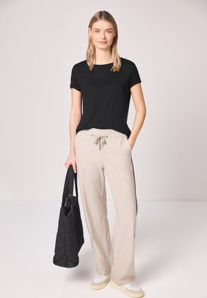 Ung kvinde stående iført en sort kortærmet top, beige bukser med snor, hvide sneakers, og holder en sort tote taske i venstre hånd.