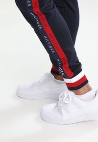 Marineblauwe joggers met rode en witte zijstrepen met de tekst "HILFIGER"; gecombineerd met witte leren sneakers met perforatiedesign.