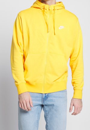 Mand iført en knaldgul Nike hættetrøje med lynlås og snore samt lyseblå jeans, stående mod en ensfarvet hvid baggrund.