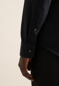 Camicia nera a maniche lunghe con polsini con bottoni, tessuto liscio e un emblema a forma di rosa sul polsino sinistro. Primo piano della camicia e della mano.