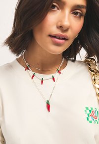 Three layered necklaces with red chili pepper pendants. Silver and gold chains paired with white beads. Cream-colored sweatshirt with logo detail.