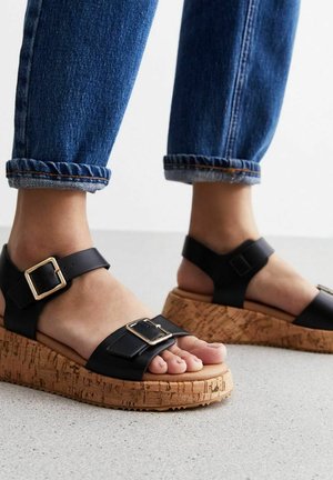 Mustat nahkaiset wedge-sandaalit, joissa on korkkipohja, leveä hihna ja säädettävä solki. Muotoilu sisältää teksturoidun korkkiyksityiskohdan.