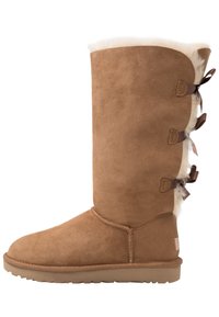 Botte haute jusqu'au genou en daim marron avec un haut en shearling blanc duveteux, des nœuds décoratifs sur le côté et une semelle en caoutchouc antidérapante.