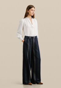 Blouse blanche à manches longues avec un col boutonné, associée à un pantalon large navy à rayures verticales et poches latérales. Chaussures marron.