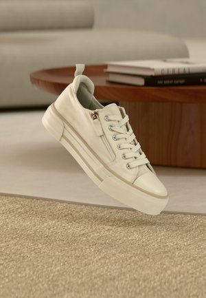 Beige afslappet sneaker med snørebånd og sidelågslynlås ophængt over et tekstureret tæppe, med et træbord og bøger i baggrunden.