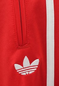 Κόκκινα αθλητικά ρούχα με φερμουάρ τσέπης, διακοσμημένα με λευκές ρίγες και λευκό κεντητό λογότυπο adidas που μοιάζει με έμβλημα τριφυλλιού.